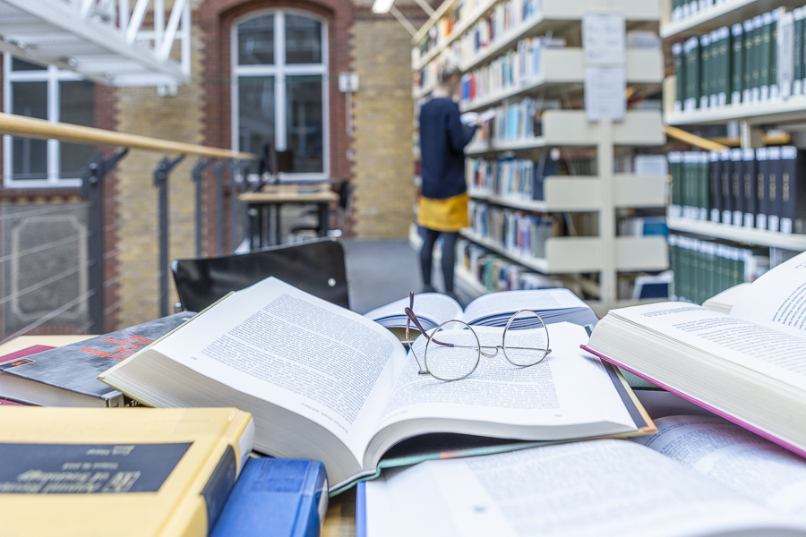 Tisch mit vielen Büchern, Brille, Frau im Hintergrund, Bibliothek